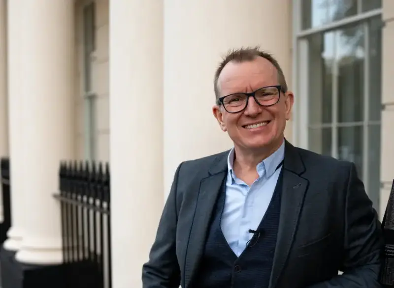 Dr Barry Oulton standing in front of a white Georgian colonnade, smiling to camera in a grey suit.