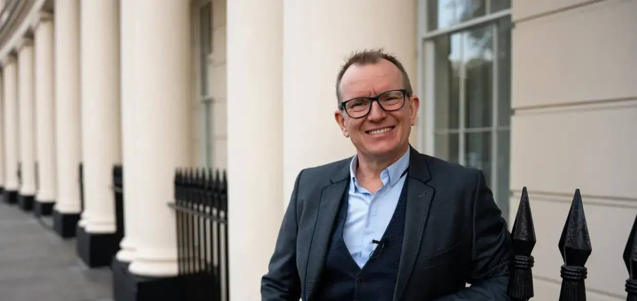 Dr Barry Oulton standing in front of a white Georgian colonnade, smiling to camera in a grey suit.