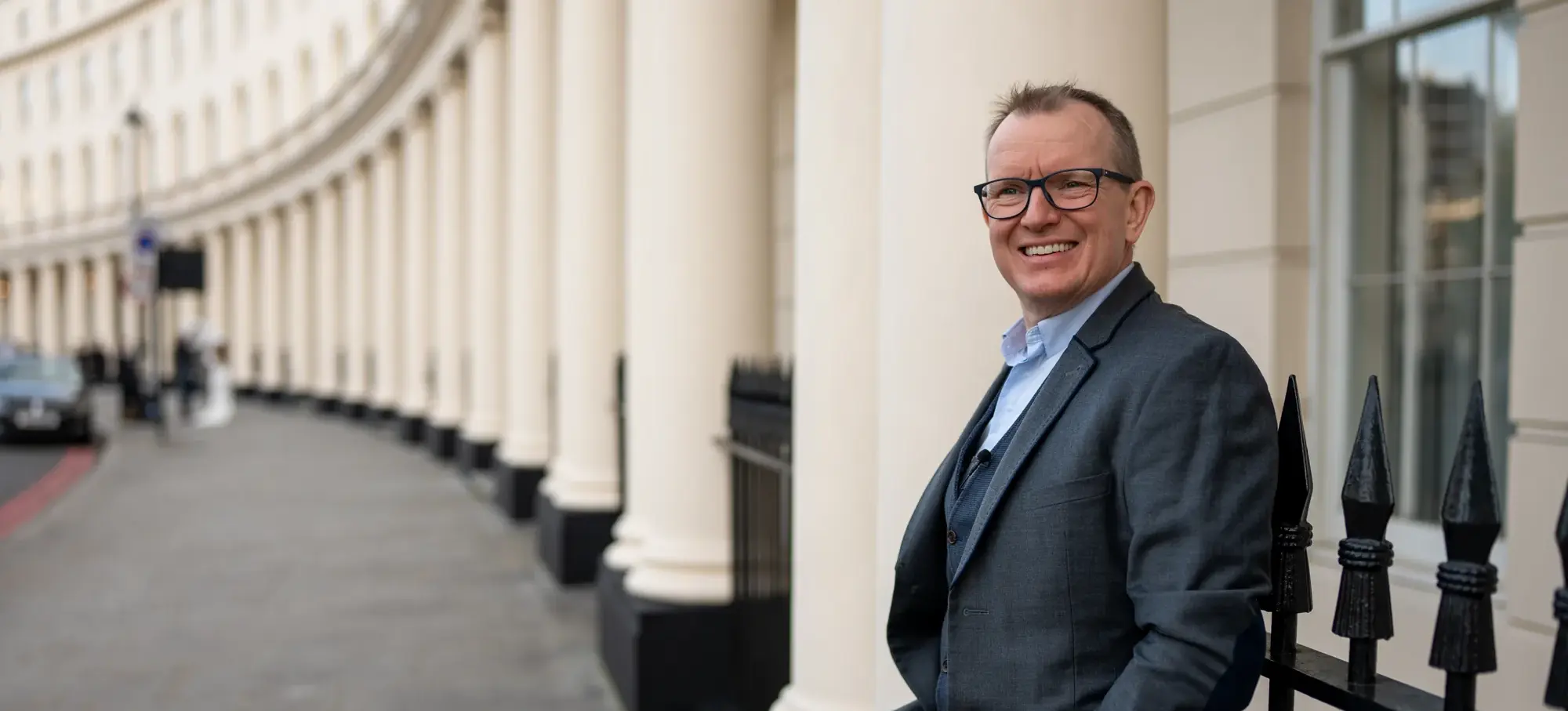 Dr Barry Oulton standing against the railings of a curved Georgian terrace, smiling to camera in a grey suit.