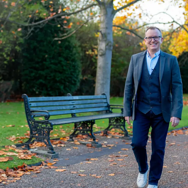 Dr Barry Oulton walking along an autumn park path past a bench, smiling to camera in a grey blazer.