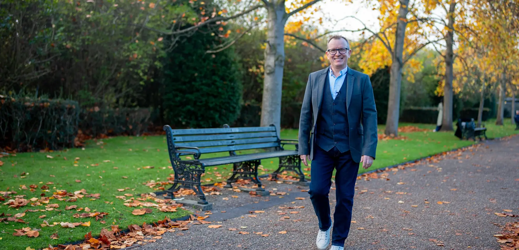 Dr Barry Oulton walking along an autumn park path past a bench, smiling to camera in a grey blazer.
